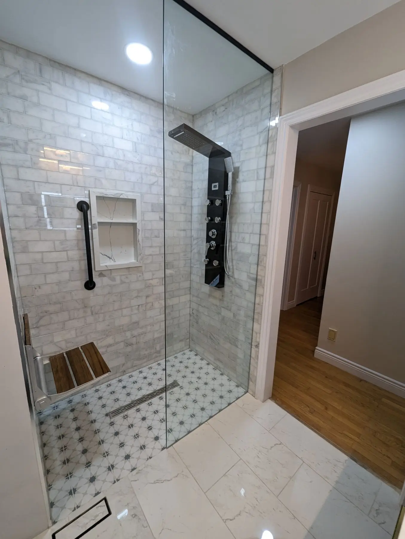 Finished — Completed walk-in shower with glass partition, marble subway tile walls, patterned floor tile, and black fixture panel.